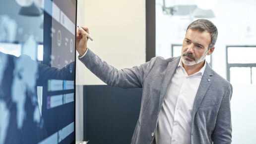 Confident male professional looking at infographic on screen in office. Mature businessman is holding with felt tip pen while contemplating. He is planning while standing in board room.